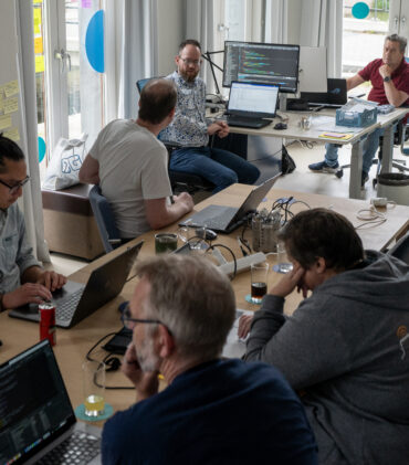 Deelnemers aan de Hackaton achter laptops aan een grote tafel en verderop in overleg aan bureau's